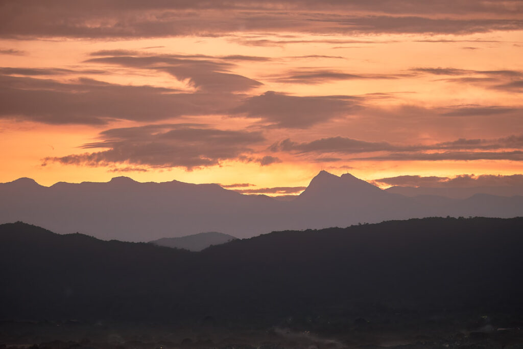 Silueta de montañas en primer plano, con una cordillera y un cielo naranja al fondo, adornado con nubes