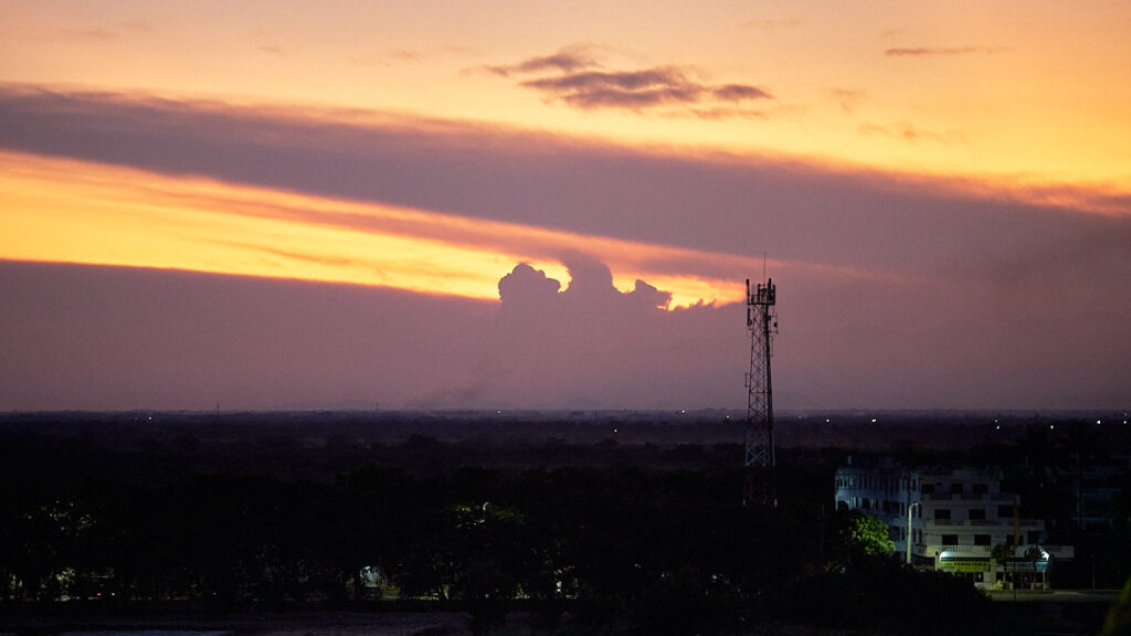 El atardecer pinta el cielo de naranja, las nubes lo cortan con armonía. Una antena de telecomunicaciones se yergue en el horizonte, su silueta se funde con las nubes, creando la ilusión de fuego emerger desde su punta. Desde mi balcón, observo este espectáculo celestial fusionado con lo urbano.